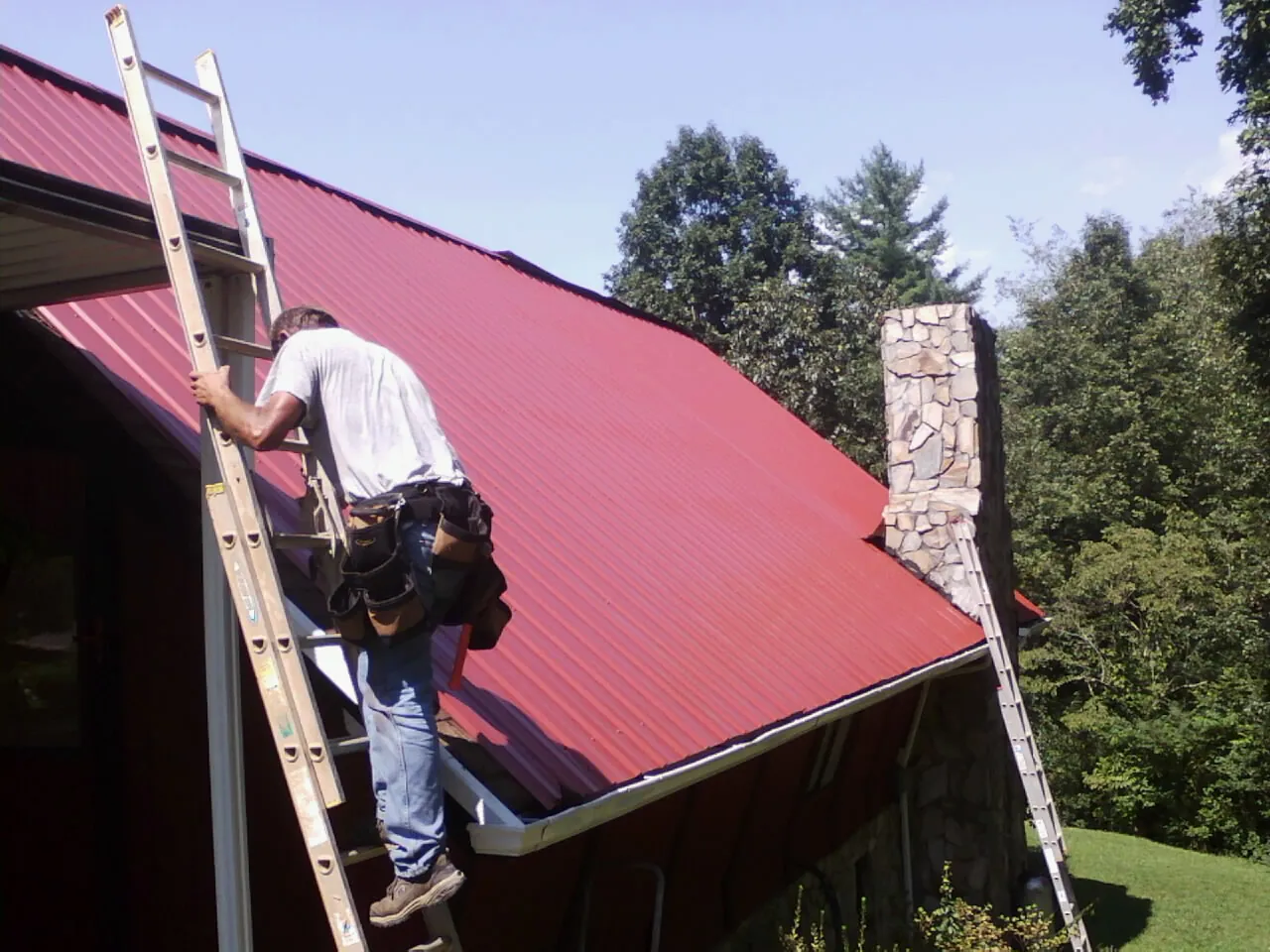 Roofer on a ladder beside a red metal roof and stone chimney — Soffit & Fascia Repair by Roofing Brothers in East Canaan